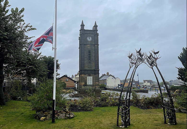 Union Jack flying at half mast in Amble. The clock tower and peace sculpture look sombre against the rainy sky